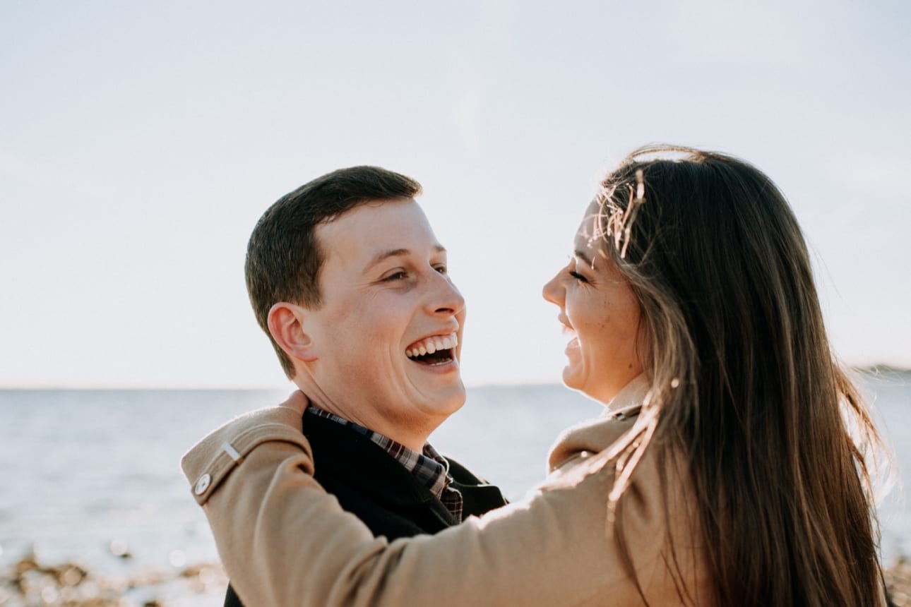 man in his 20s laughs as his fiancee hugs him and smiles at the beach in connecticut during an early spring engagement session with Kristyn Miller Photography