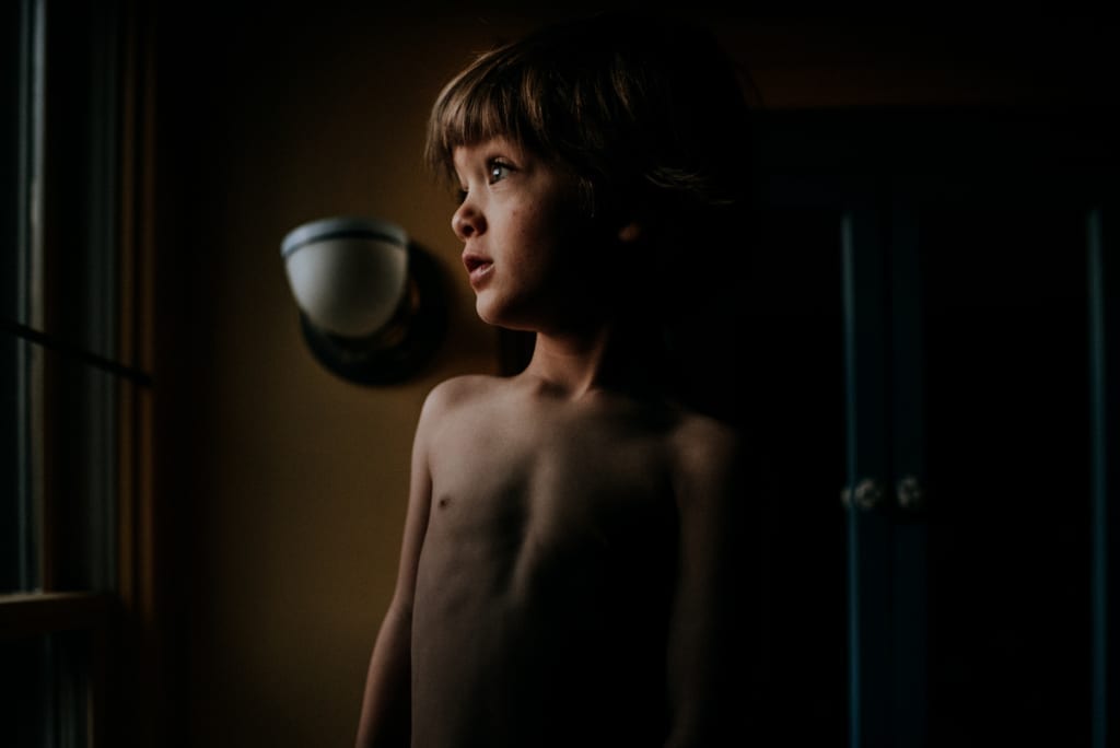 little shirtless caucasian boy stands in a dark room looking toward a window into the unknown. Raising kids is kind of like riding the lion. Image by Kristyn Miller Photography.