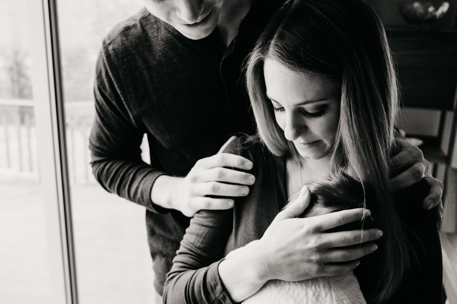 a new mom holds her newborn to her chest with her husband hugging her and smiling down at them during an newborn session at home with Kristyn Miller Photography