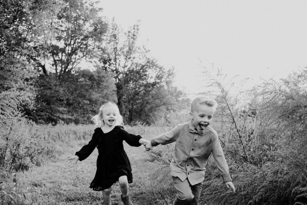 a happy image by Kristyn Miller Photography where a little blonde girl and her brother hold hands as they run through the forest smiling