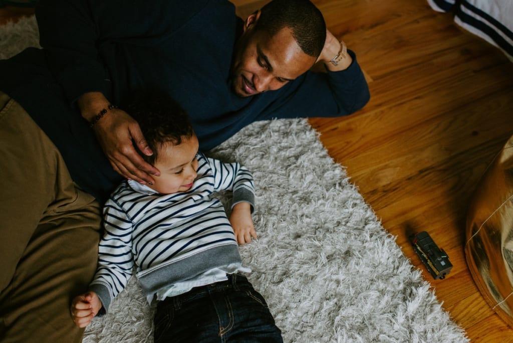 a black man lays on the floor with his toddler son and they smile and he holds his son's head