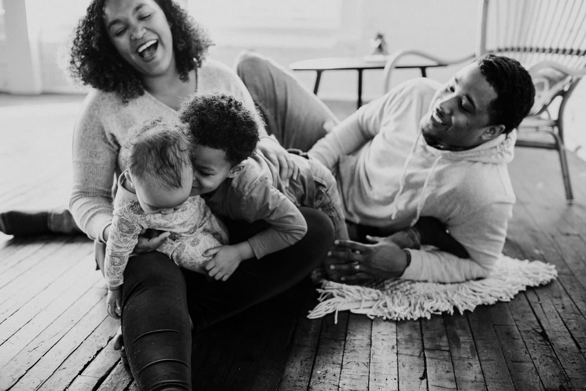 a young black family with a toddler son and a baby girl sit on the the floor and share a laugh as the brother gives his sister kisses in a beautiful display of cultivating kindness by Kristyn Miller
