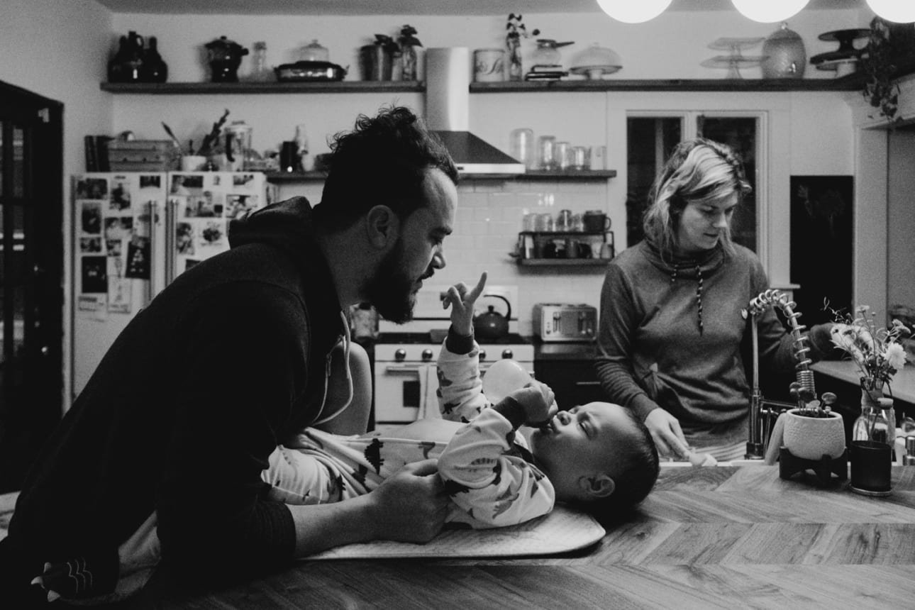 a dad plays with his baby son on the counter in the kitchen while mom prepares coffee in this in-home session with Kristyn miller photography