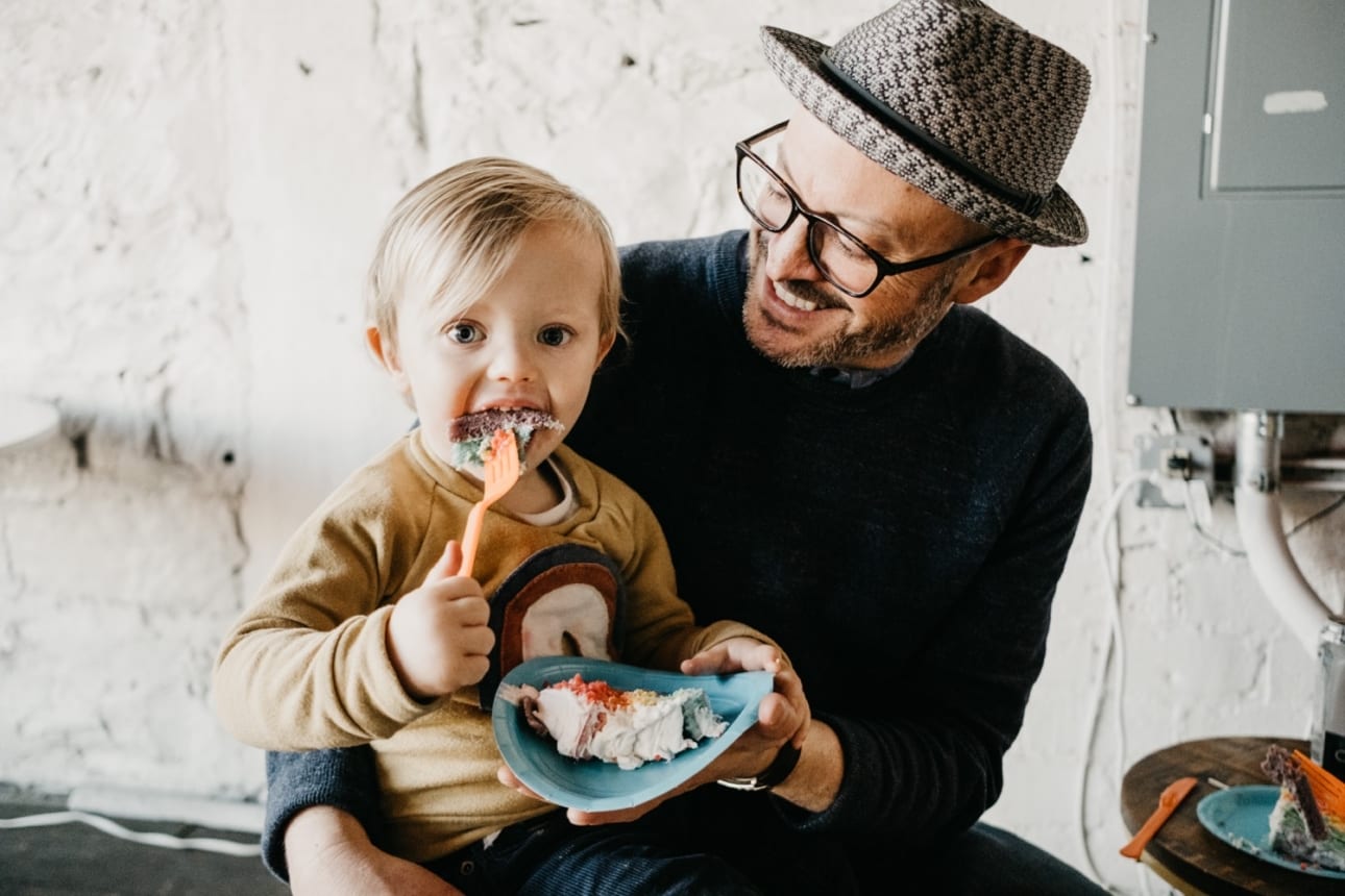 little boy eats rainbow cake at his 2nd birthday party while sitting on his dad's lap. Candid documentary images by Kristyn Miller Photography