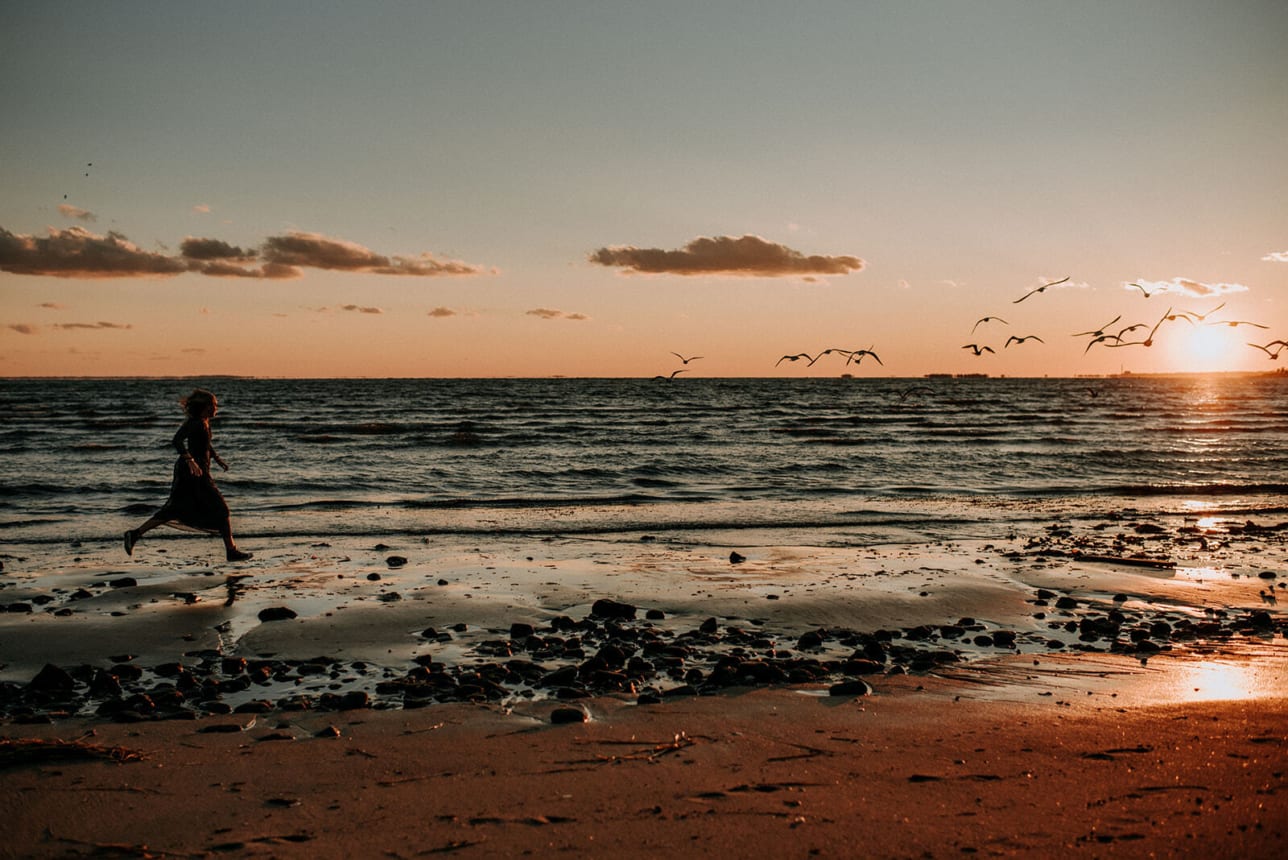 woman runs along the beach as the sun sets and seaguls fly around over the water in the distance. Image by Kristyn Miller Photography