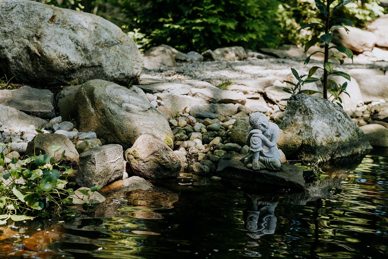 a buddha statue rests with hands on knees and head on hands on a flat rock in a beautiful and peaceful garden and pond setting
